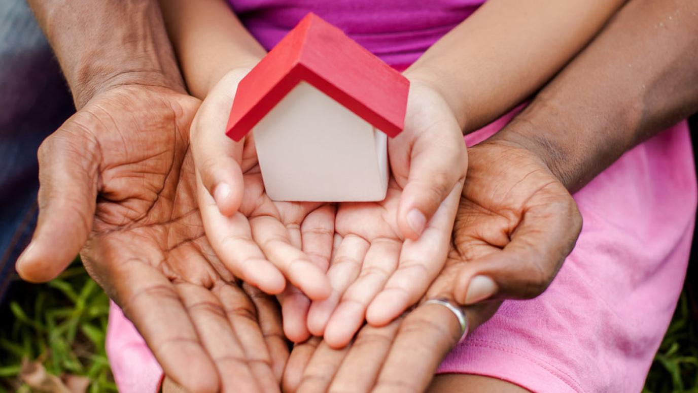 A house rests in the open palms of a child. The child's hands rest in the open palms of an adult. 