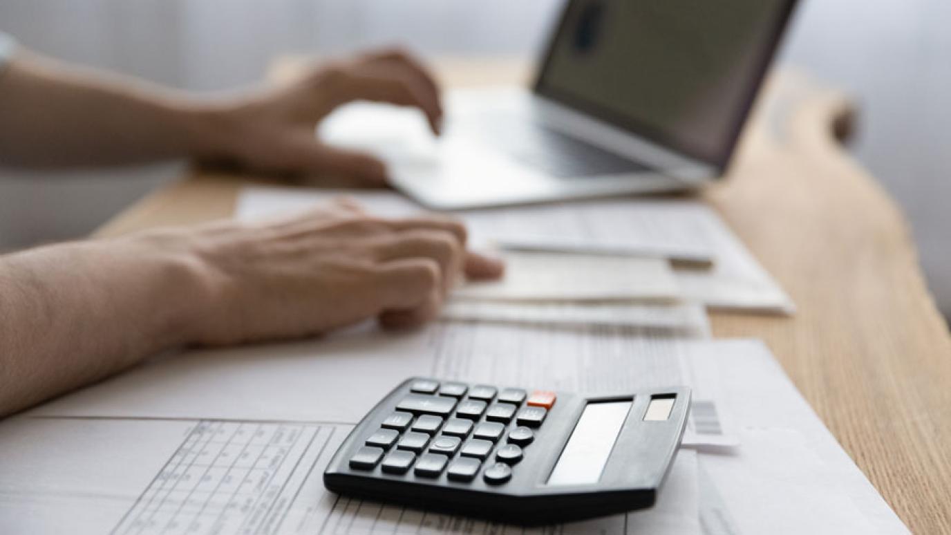 Close up of young mans hands working with invoices using a laptop. Focus on electronic calculator lying on desk.