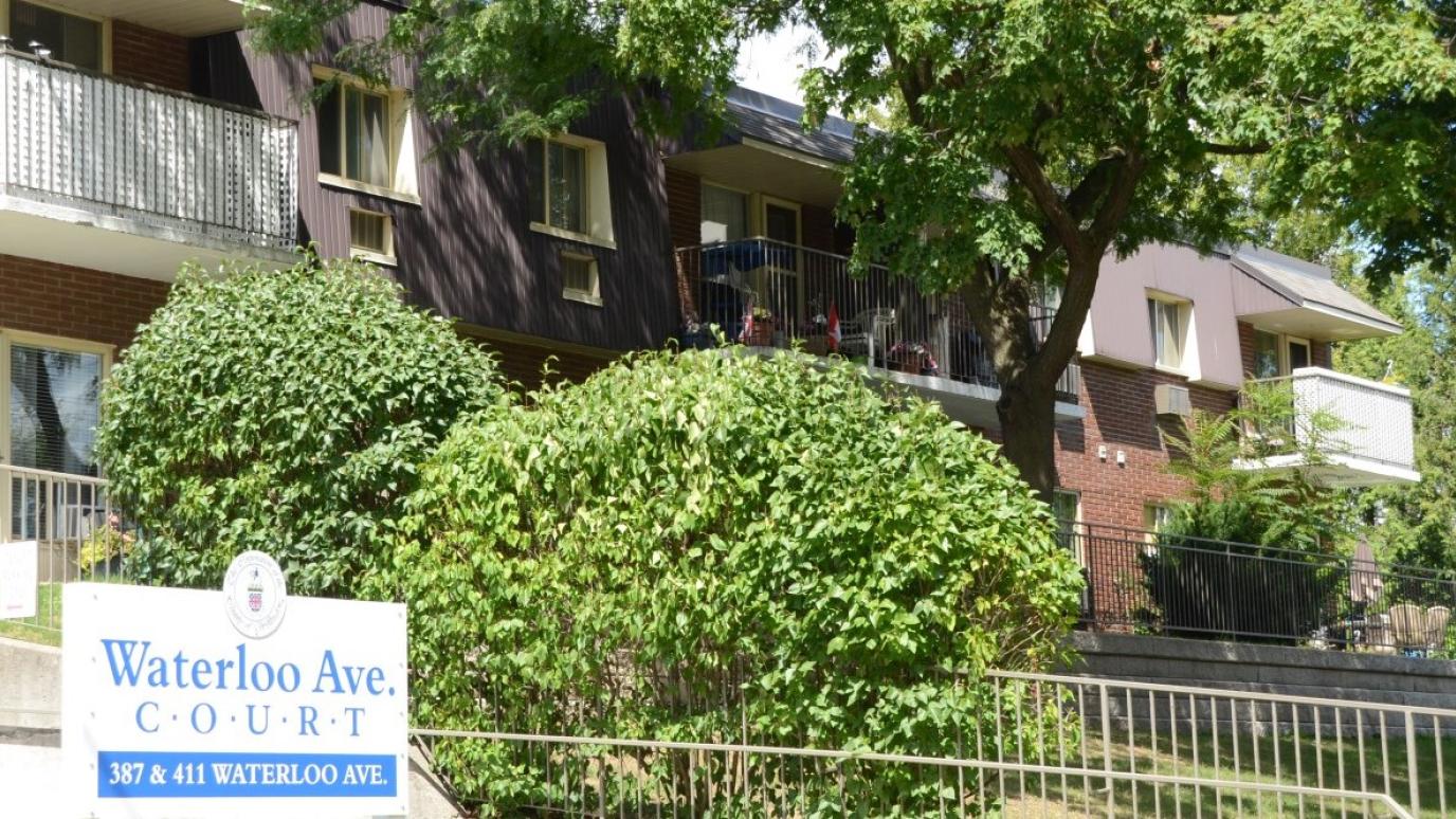 Exterior of 387-411 Waterloo Avenue, Guelph. It is a two-storey apartment building elevated from the street with trees in front.