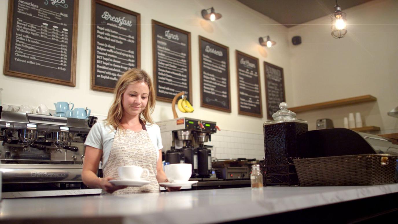 Barrista smiles while holding two coffees behind a counter with coffee machines in the background.
