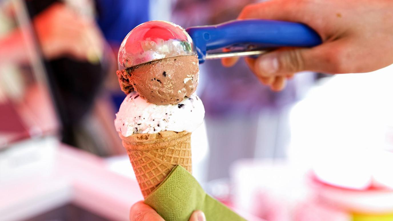 A blue ice cream scooper adds a scoop of chocolate ice cream onto waffle cone.