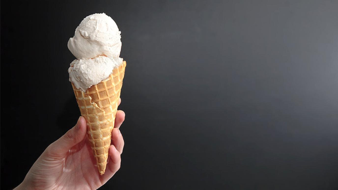 Hand holds up a waffle cone of two scoops of vanilla ice cream in front of a black background.