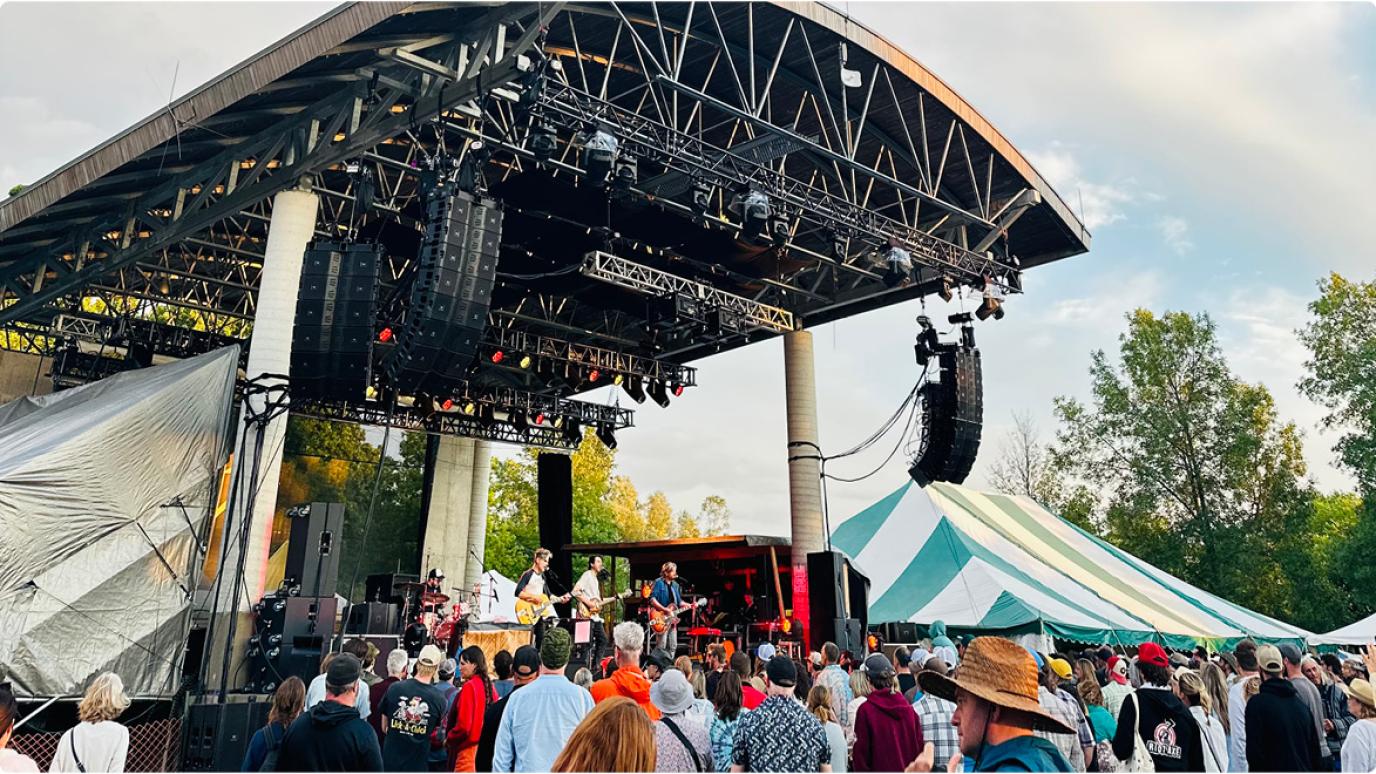 Photograph of a band performing on the main stage at the Hillside Festival with crowds of people in front, stripped tents in the background, and blue skies above. 
