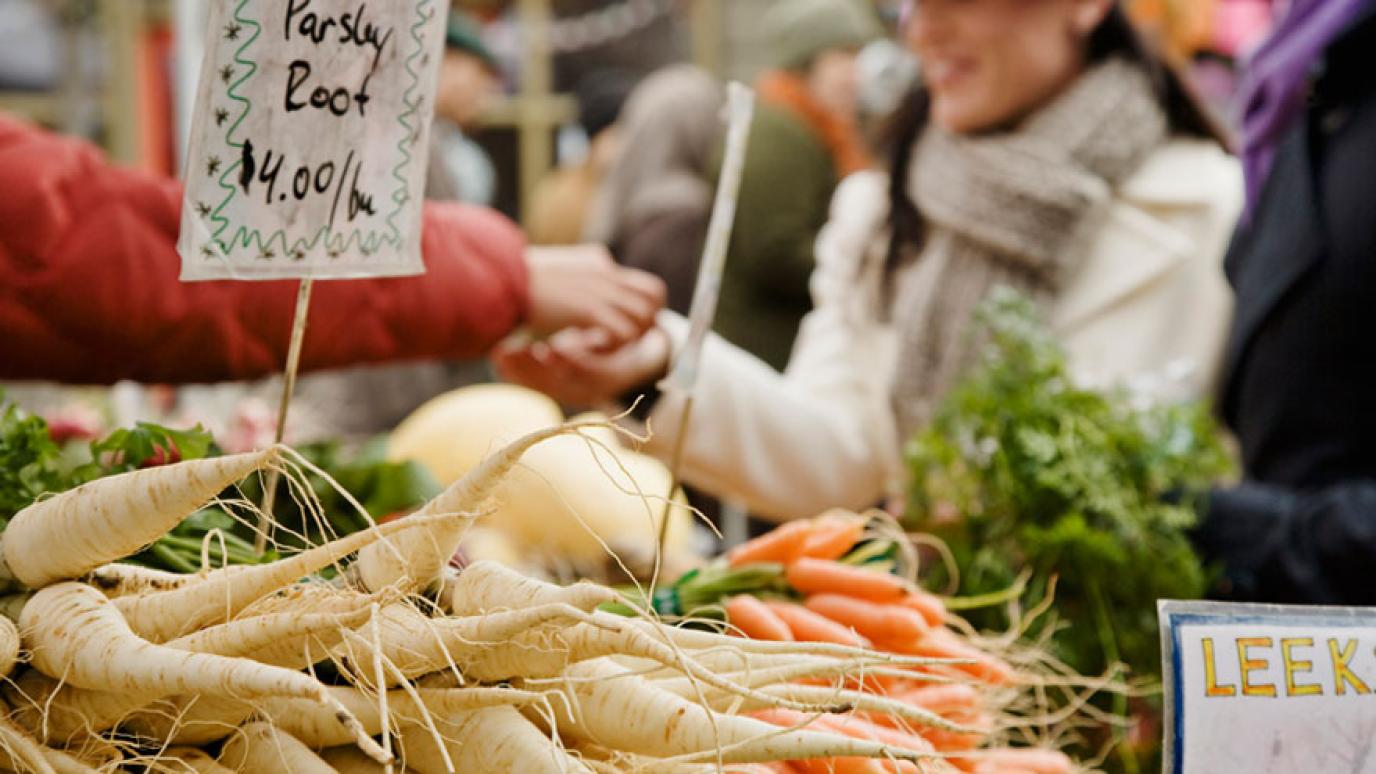 A couple dressed in winter attire buy parsnips and carrots at an indoor farmer's market stand