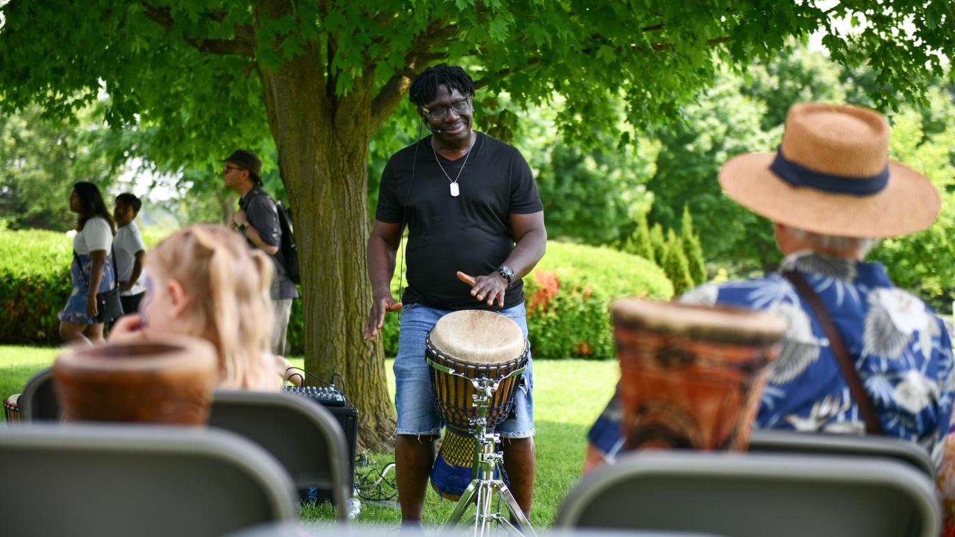 A smiling man leads an outdoor African drumming circle under the shade of a large tree, surrounded by seated attendees holding djembes.