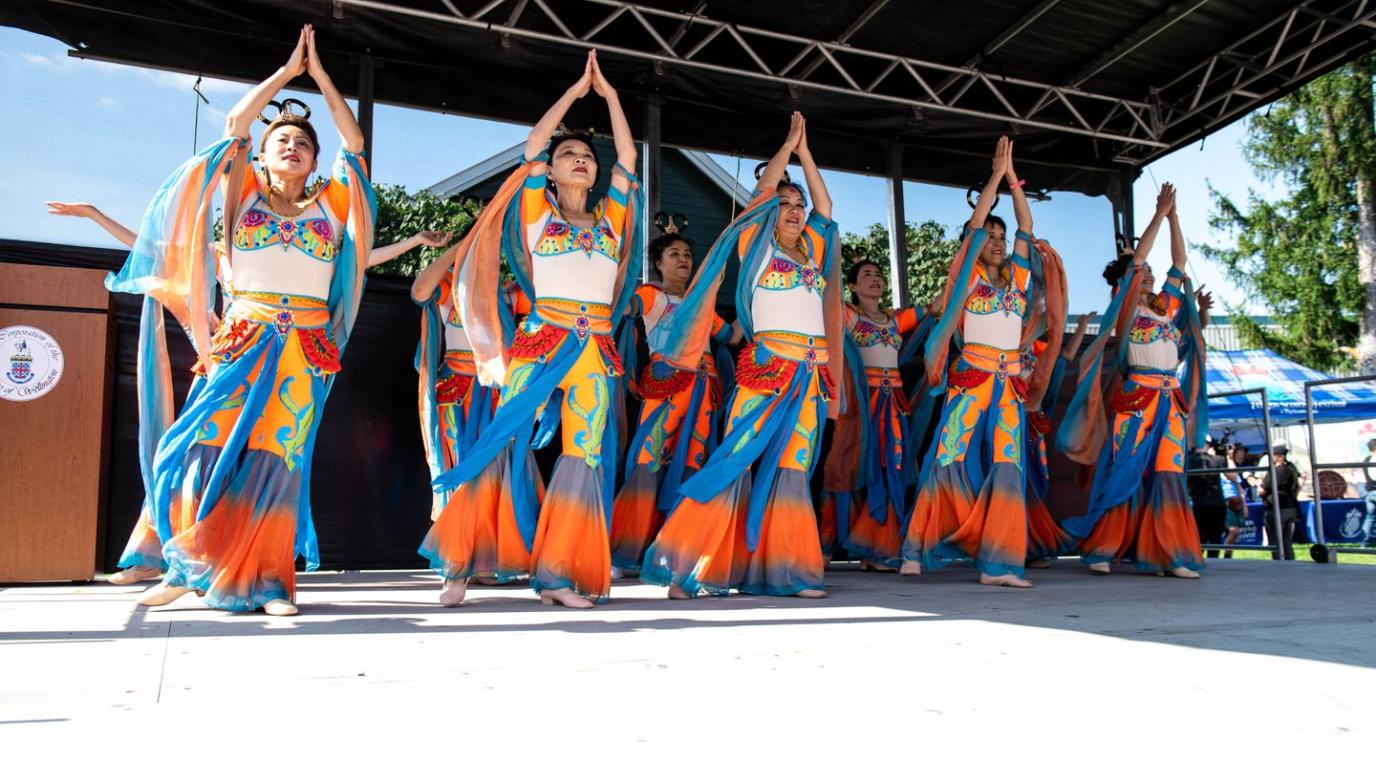 A vibrant group of dancers in coordinated orange, blue, and gold costumes perform a traditional dance on an outdoor stage, hands raised in unison.