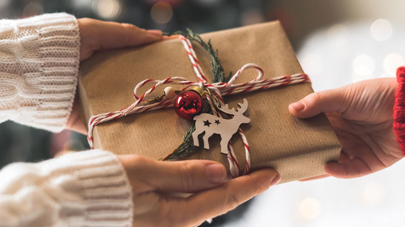 A woman's hands pass a christmas present wrapped in kraft paper with ribbon and a wooden reindeer on top, received by a childs hand.