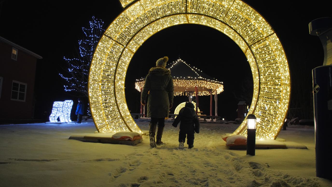 A mother and child walking towards a large, illuminated arch decoration on a snowy night, with a distant gazebo visible through the arch.
