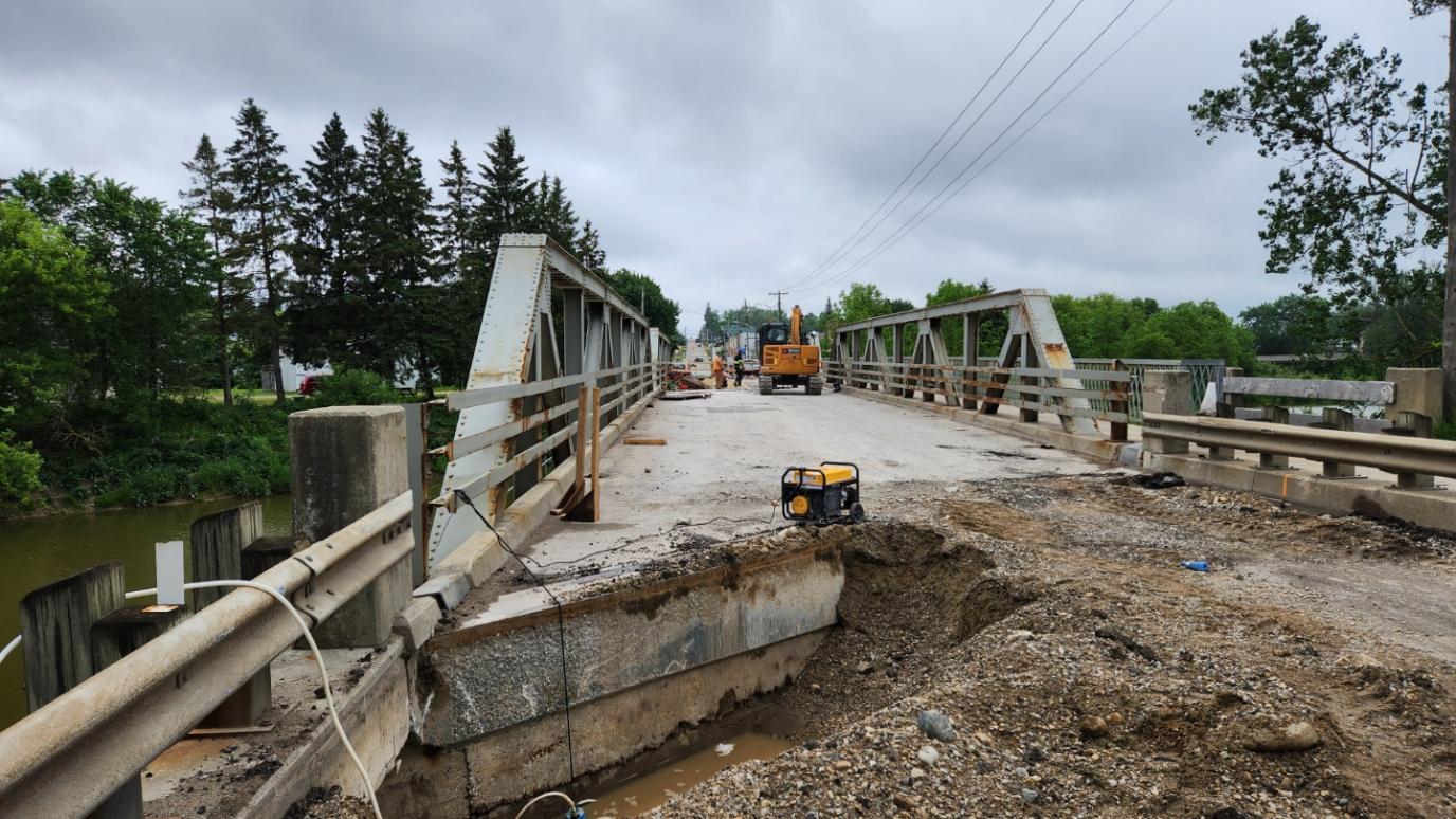 Damaged bridge with a large hole in the concrete surface under cloudy skies and construction equipment in the background.