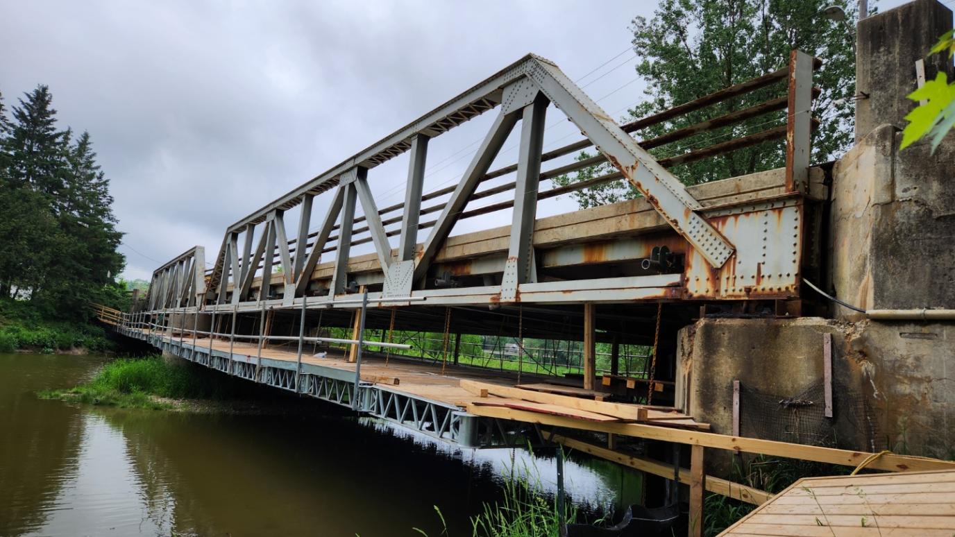 Rusty metal truss bridge spanning a calm river under a cloudy sky with green trees along the banks.