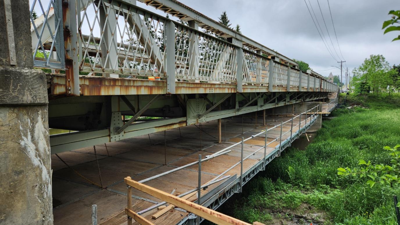 Rusty metal truss bridge with a wooden plateformunderneath, surrounded by green vegetation and cloudy sky.