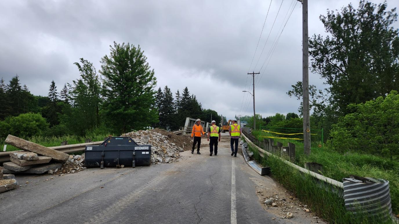 Three workers in safety vests walk on a road blocked by construction debris and equipment under a cloudy sky. 