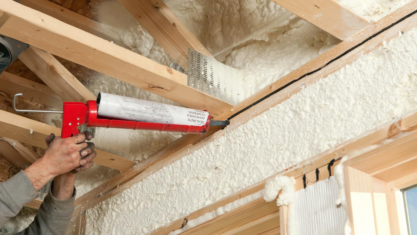 A construction worker is applying sealant to a gap between two window header boards at a house construction site. 