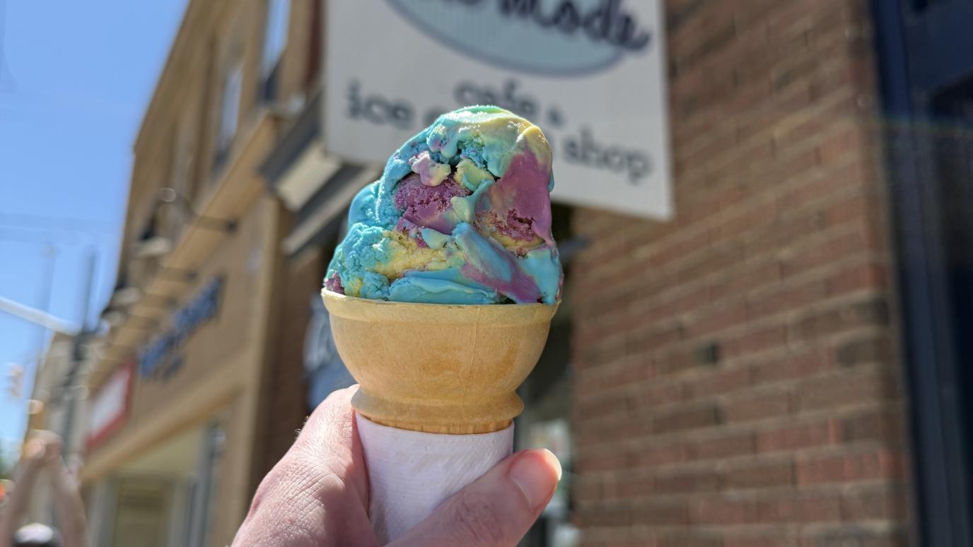 hand holding colourful scooped ice cream in a regular cone in front of a store sign