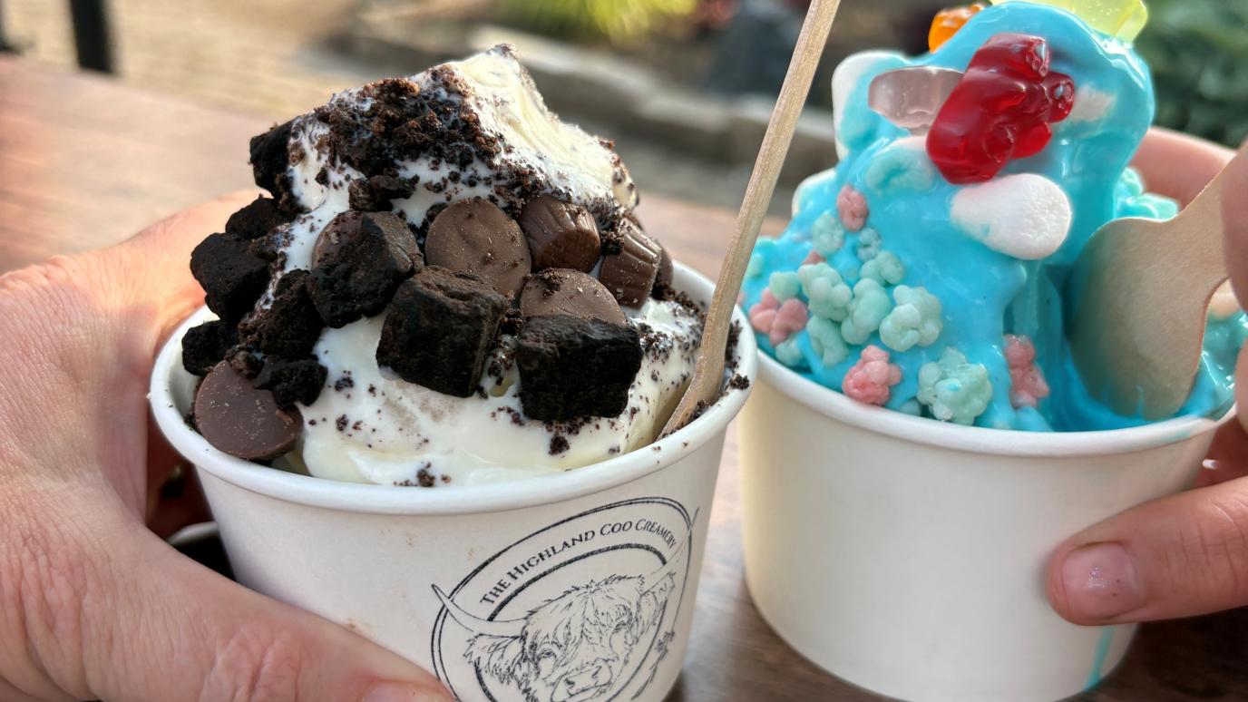Two hands holding paper cups of ice cream outdoors—one vanilla topped with brownie bites and cookies, and the other bright blue with colorful candy toppings