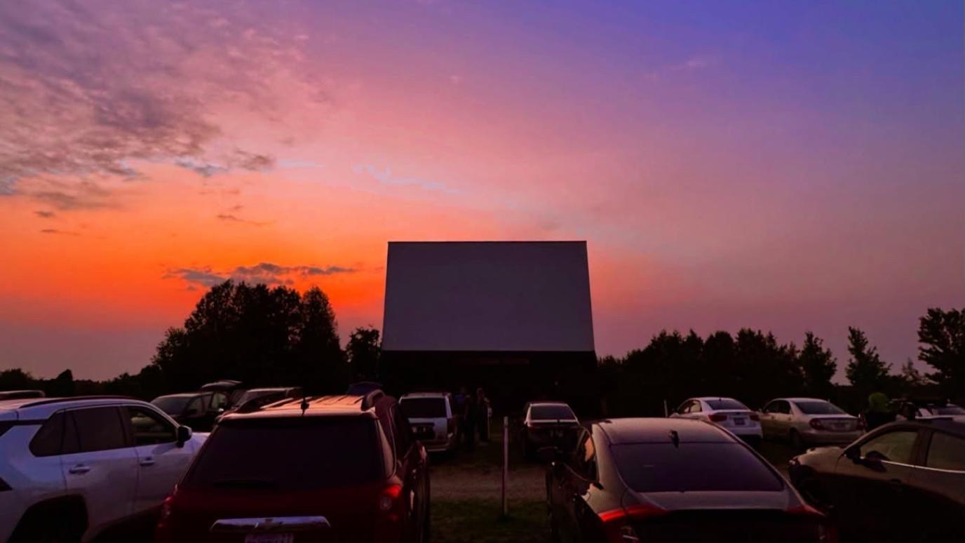 Drive-in movie theater at sunset with parked cars silhouetted against a brilliant orange and purple dusk sky.