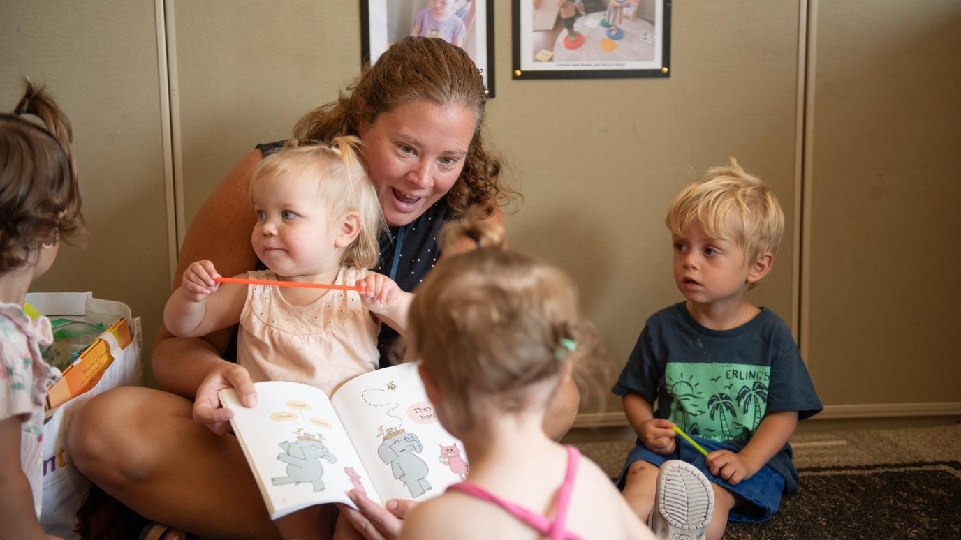 An ECE sits reading a books to a bunch of kids sitting around her. 