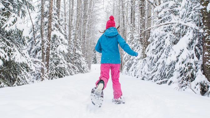 A young girl runs along a snowy trail lined with trees wearing snowshoes and bright winter gear.
