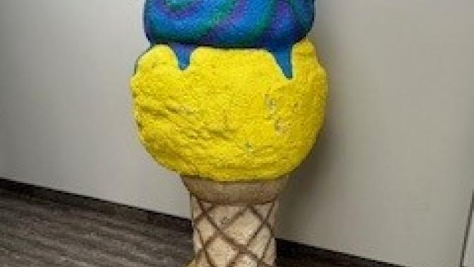 Large sculpture of a three-scoop ice cream cone with yellow, blue-purple, and red scoops, displayed indoors against a plain wall on a wood-style floor.