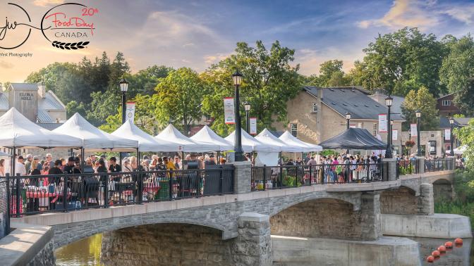 A wide-angle shot of the pedestrian bridge in Elora, which is lined with white tents and people crossing the bridge.