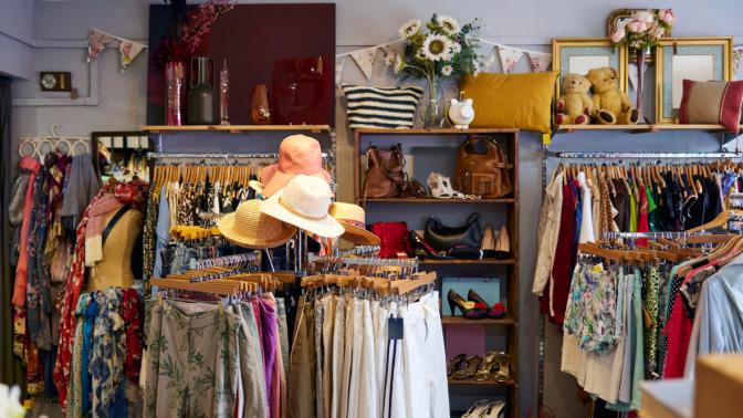A cozy and colorful thrift store interior with neatly arranged racks of second-hand clothing, hats, handbags, and shoes. The store features decorative items such as pillows, flowers, framed mirrors, and plush teddy bears on upper shelves, creating a welcoming and eclectic atmosphere. A display case on the left showcases jewelry and accessories.