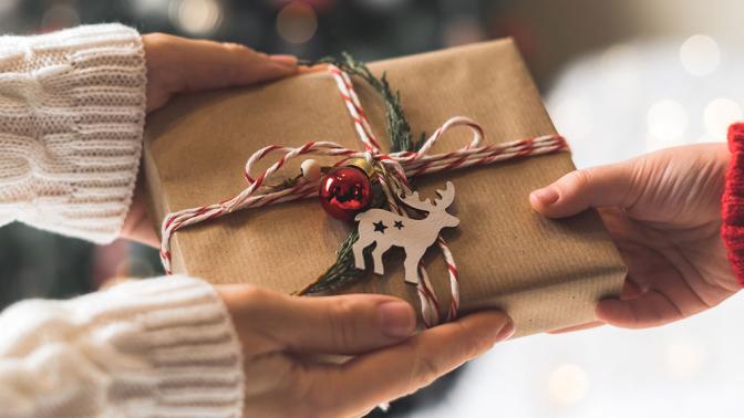 A woman's hands pass a christmas present wrapped in kraft paper with ribbon and a wooden reindeer on top, received by a childs hand.