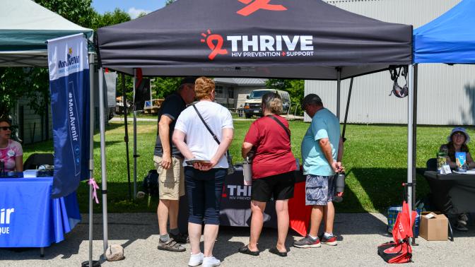 Several people gather under a black "Thrive HIV Prevention and Support" canopy tent at the Multicultural Festival on a sunny day.