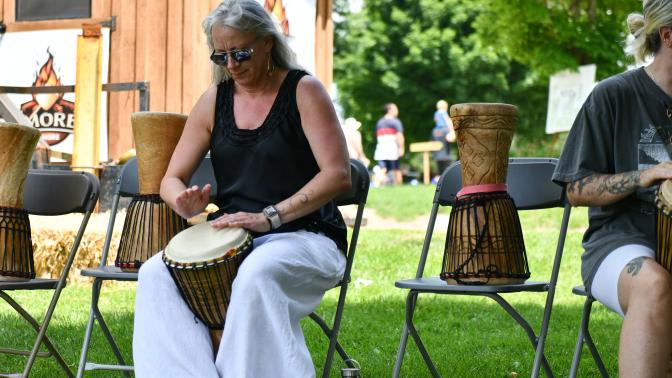 A woman in a black shirt and flowy white pants sits on a folding chair in a grassy area, playing a drum during the Multicultural Festival. 