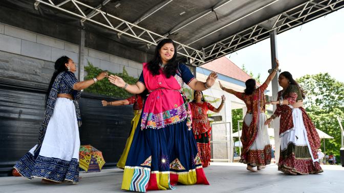 Five women in colorful, traditional South Asian attire perform a choreographed dance on an outdoor covered stage.