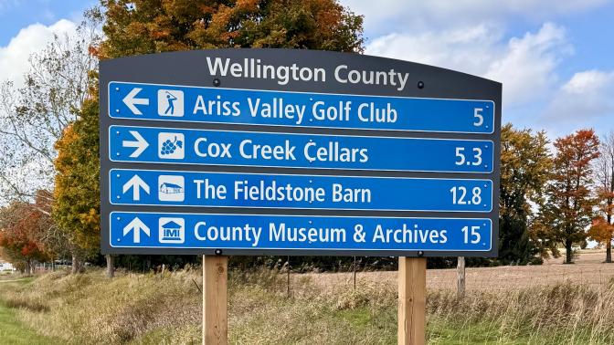 Wellington County directional tourism signage. The four sign blades feature the following businesses: Ariss Valley Golf Club, Cox Creek Cellars, The Fieldstone Barn, and County Museum and Archives.