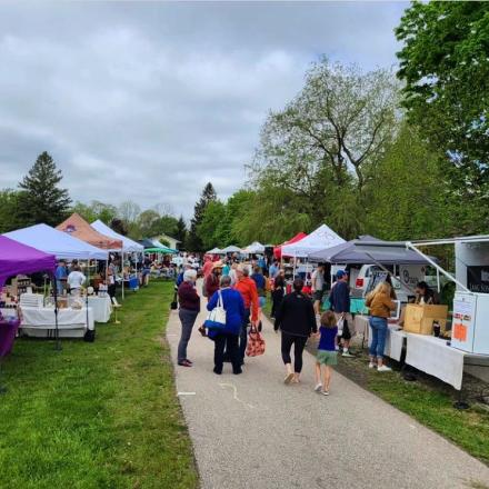 Groups of people walking along a trail surrounded by Farmers' Market vendor tents.