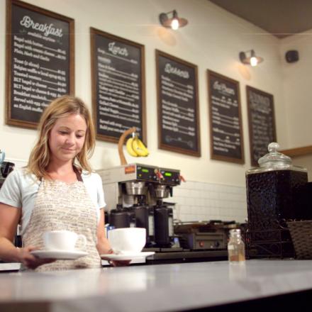 Barrista smiles while holding two coffees behind a counter with coffee machines in the background.
