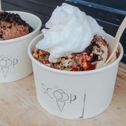 Two cups of ice cream with the Scoop logo on the front; one full of chocolate ice cream with cookie crumbs on top, the other filled with a vanilla and whipped cream toppings. 