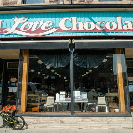 The outside of a storefront with a large blue, white and red sign that reads “I Love Chocolate and Gifts” above a double window, with a bike outside of it.