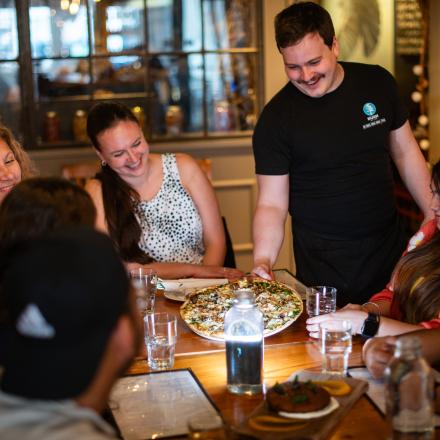 people sitting around table smiling while being served pizza