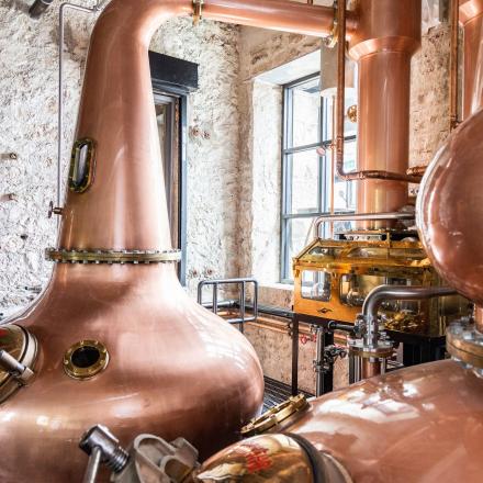 Two large, polished copper stills, likely for distilling spirits, dominate the frame. They sit within an industrial-style setting with exposed brick walls and a window, suggesting a distillery.