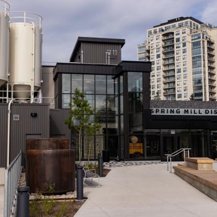 A modern distillery building with large silos on the left and a tall residential building in the background.