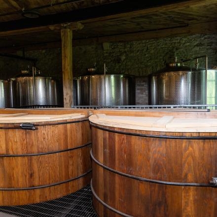 Two large wooden vats with lids are in the foreground, with several smaller stainless steel tanks visible in the background, all within an industrial setting.