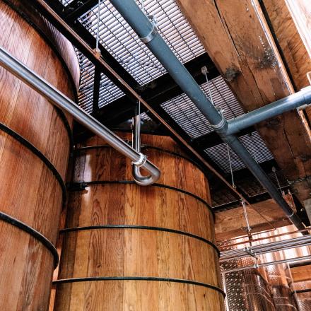 A low-angle shot of large wooden fermentation tanks in a distillery, with industrial grating and pipes above.
