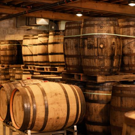 A large, dimly lit room is filled with numerous wooden barrels, stacked on pallets in a whiskey-aging cellar.