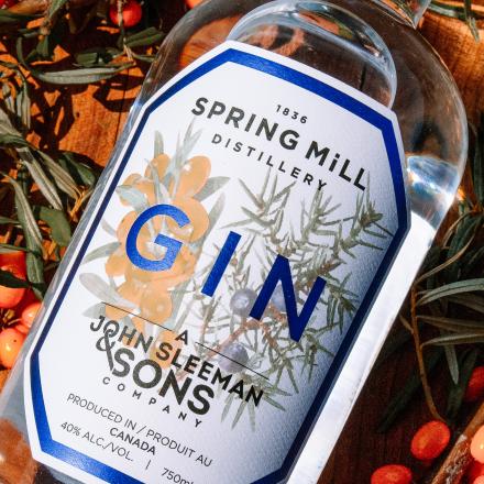 A close-up of a clear glass bottle of Spring Mill Distillery Gin, with a white and blue label, resting on a bed of green foliage and small red and orange berries.