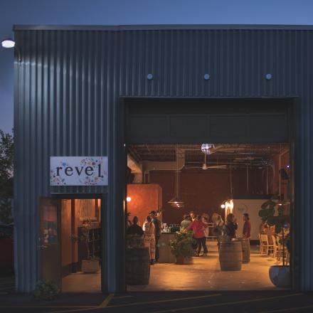 Evening view of an open industrial-style cidery with people gathered inside under warm lighting; a sign reading "revel" is mounted beside the entrance.