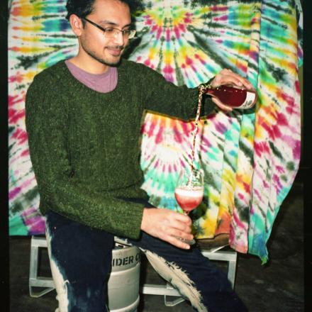 Person in a green sweater sits on a keg, pouring a drink dramatically between two glasses in front of a colourful tie-dye backdrop.