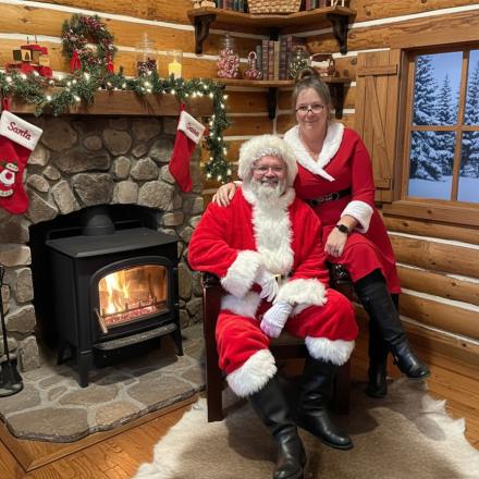 santa claus and a woman in a red suit sitting by a wood stove in a log cabin