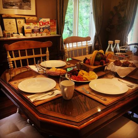 A wooden dining table set for breakfast with white plates, fruit, bread, and juice, located in a sunlit room with a window and a sideboard displaying cereal boxes.