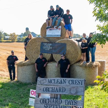 A group of people posing on a pyramid of large round hay bales behind a wooden pallet sign advertising "McLean Crest Orchard" and an event in September