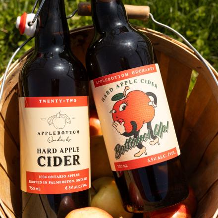 Two swing-top bottles of hard apple cider in a wooden basket filled with fresh apples.
