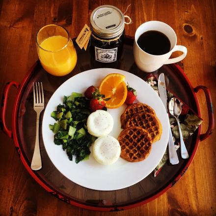 A breakfast tray featuring a plate with poached eggs, mini waffles, greens, and fruit, served with orange juice, coffee, and a jar of preserves.