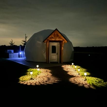 An illuminated dome tent with a wooden entryway frame at night, featuring a pathway lit by patterned outdoor lights.
