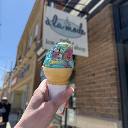 hand holding colourful scooped ice cream in a regular cone in front of a store sign